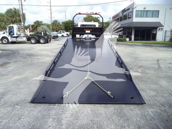 Flatbed tow truck bed with chains and hooks, Peterbilt 337 in background, dealership building on right.