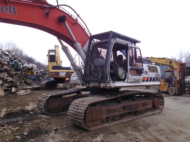 Excavator with long orange boom and tracked undercarriage in a scrapyard; cabin with metal guard, Caterpillar logos, and other heavy equipment in background.