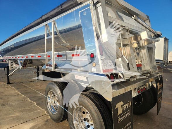 Dump trailer with shiny metal sides and dual rear wheels showing chrome rims, hydraulic tailgate, and reflective markings. Fleet logo and equipment tag visible.