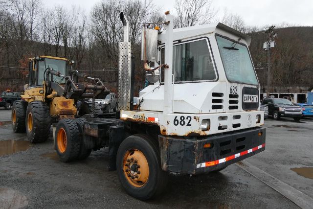 Heavy-duty TJ5000 yard truck with white cab and yellow wheels, capacity label '682' on front, connected to a heavy machinery trailer, overcast outdoor lot.