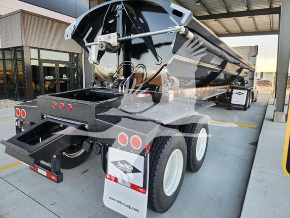 Smithco 2025 dump trailer shown from rear-left side with tri-axle wheels, black exterior, hydraulic dump mechanism, and rear tailgate open area.