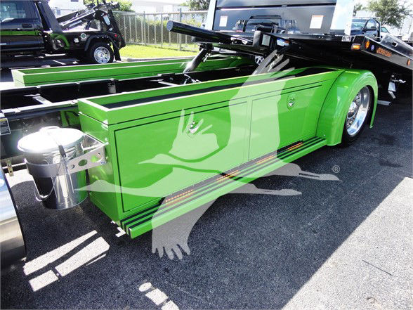 Green Peterbilt truck storage bed with glossy finish, side toolboxes, and chrome fuel cap; trailer hitch visible; other trucks in background.
