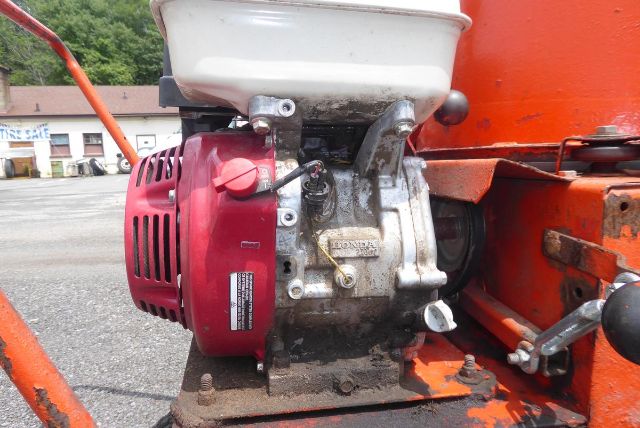 Small gasoline engine mounted on orange agricultural machinery, with white plastic fuel tank above and red recoil housing; visible label on engine side.