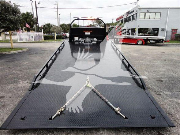 Tow truck flatbed with hydraulic winch and chain anchors on black diamond-plate deck. Tow equipment includes side rails and a rear winch visible at cab.