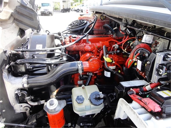Freightliner M2 106 engine bay showing red diesel engine, coolant reservoir, battery, hoses, and various components.
