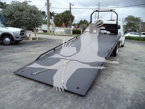 Tow truck with a flatbed in lowered position. Flatbed surface shows chains and hooks. Background includes other trucks and a parking lot.