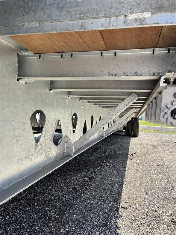 Underbelly of a 2024 Manac steel drop deck trailer showing structural beams, perforated side frame with circular cutouts, and wheel in distance.