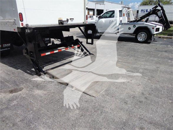 Box truck rear view showing a liftgate deployed and a second white truck with a crane in the background.