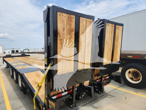 Behnke flatbed trailer with extendable ramps folded up, wooden deck, dual rear ramps, yellow strap, black frame, and triple-axle configuration visible.