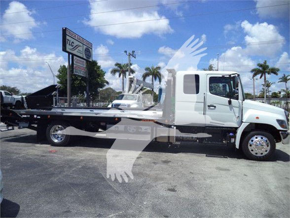 Flatbed tow truck in a dealership lot, white cab with long flatbed, palm trees in background, clear sky.
