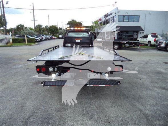 Tow truck with flatbed at rear showing license-plate region, rear lights, and reflective safety lights; flatbed surface with slight incline; dealership lot in background.