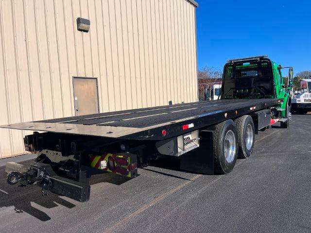 Flatbed tow truck with a long black trailer on a Freightliner M2 106 chassis, green cab visible, triple rear axles, equipment and hookups under trailer.
