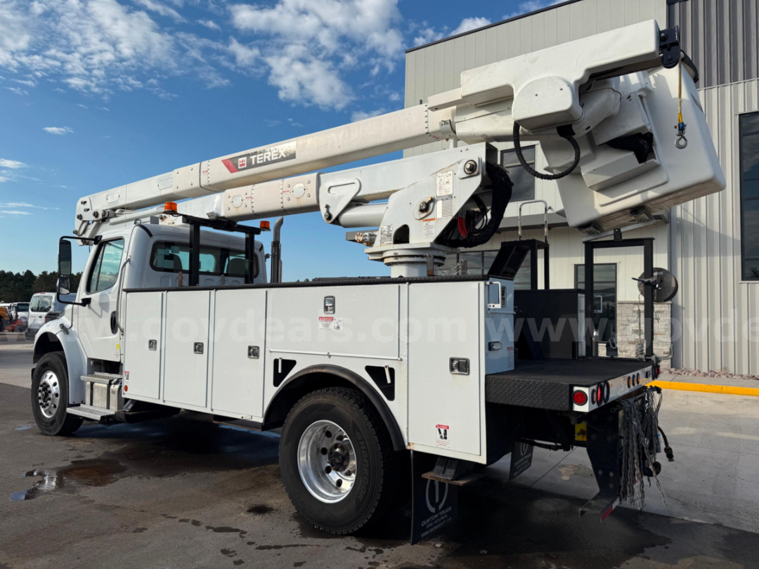 Truck-mounted bucket crane with white utility body and Terex boom, side compartments visible, rear platform and stabilizers, Freightliner chassis, clear blue sky.