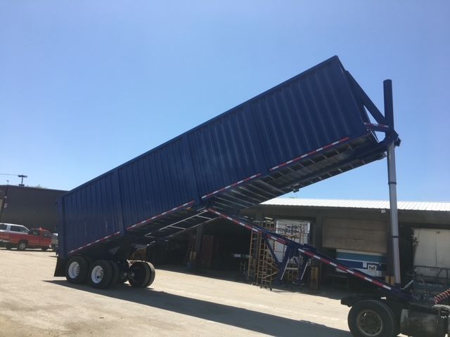 Trailer showing a blue 40 ft square dump trailer raised on its hydraulic hoist, triple axle with dual tires, in a yard with a building in the background.