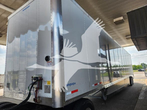 Wabash aluminum trailer under a canopy showing storage access and electrical hookup on the front corner.