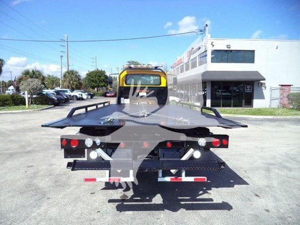 Back of a tow truck with a flatbed. Tail lights, license-plate area, and hydraulic components visible. Foreground shows the rear of the bed with wheel tracks and support arms.