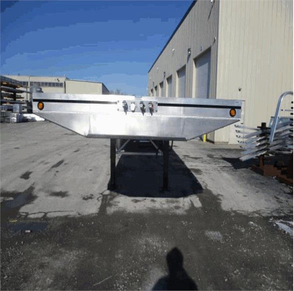 Tri-axle flatbed trailer with aluminum beam and legs, facing front with stakes and reflectors.