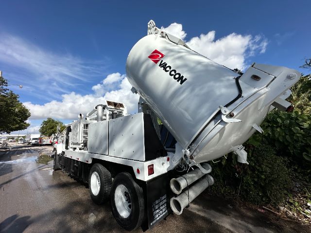 Vac-Con street-angled image of a large white vacuum truck with a tilted cylindrical tank labeled VAC-CON, multiple hoses, and a chrome exhaust area.