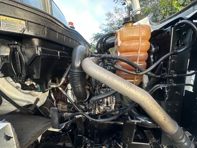 View of engine bay in a 2016 International 7400 showing coolant reservoir, hoses, and air intake. Orange coolant/overflow reservoir at back with black caps. Various black hoses and metal components.