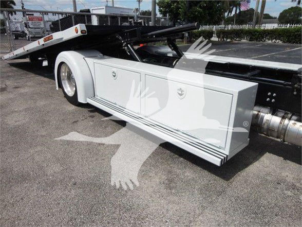 Tractor-trailer tractor unit side view showing stainless toolbox and landing gear with flatbed trailer in background.