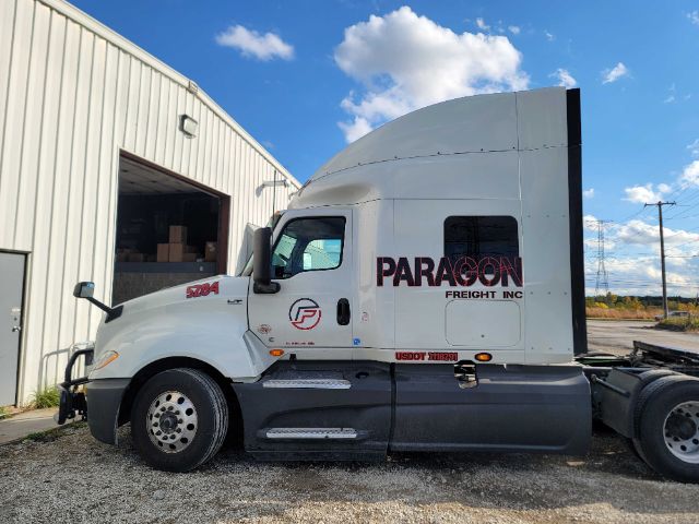 Truck cab side view showing PARAGON FREIGHT INC logo and S90? text on white sleeper cab, International LT625 model badge visible, blue sky and warehouse in background.