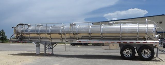 Aluminum vacuum tank trailer mounted on dual-axle trailer with cylindrical tank and manways, connected piping and railing along top.