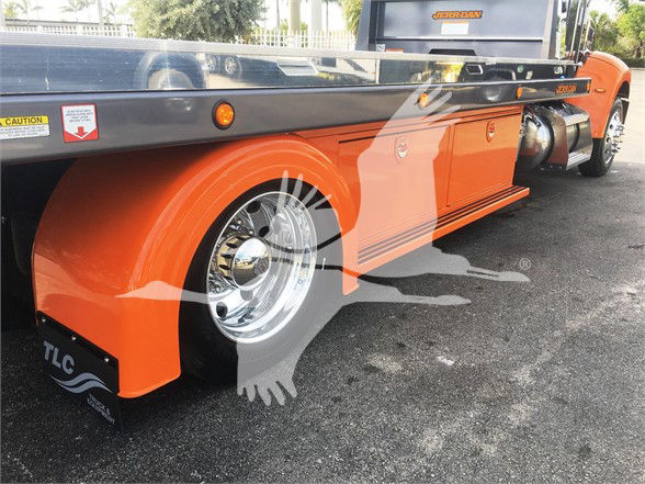 Peterbilt 337 tow truck side view showing orange fender and chrome wheel with TLC mudflap, black deck, and orange cab in background.