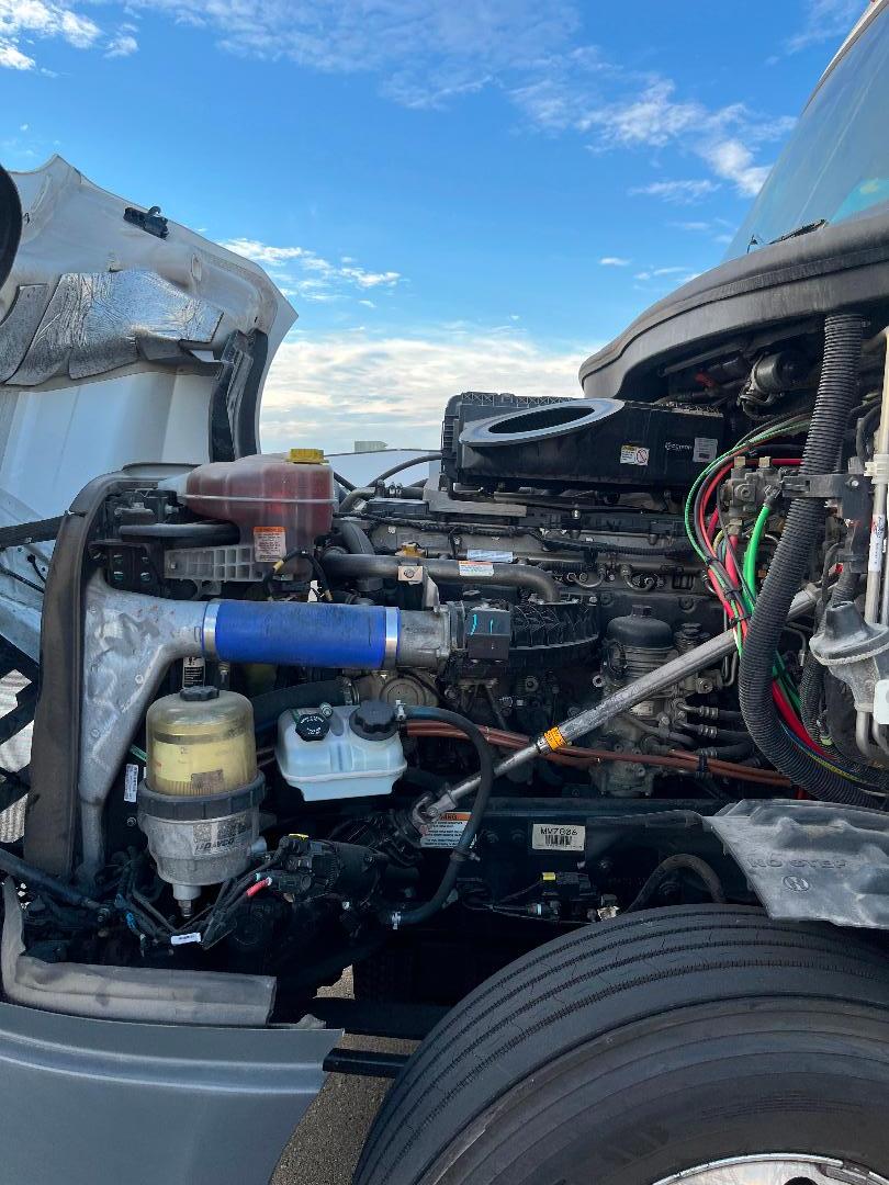 Freightliner Cascadia engine bay showing coolant reservoir, blue silicone hose, various fluid reservoirs, and wiring harness in a truck engine compartment.