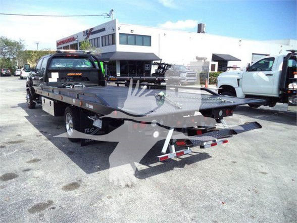 Towards rear view of a black tow truck flatbed with a white commercial building in background. License plate area and stickers visible on the cab window.