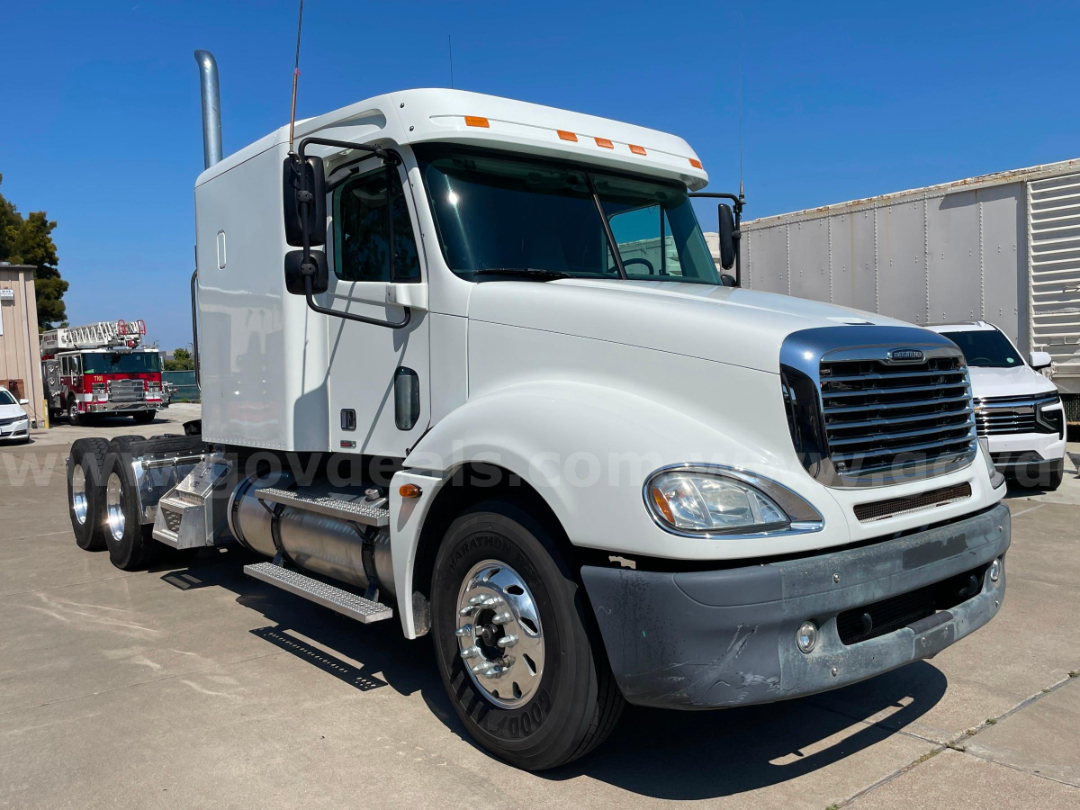 Truck cab and chassis showing Freightliner badge, front grille, and side steps with dual rear axles. Windshield mirrors and chrome wheels visible.