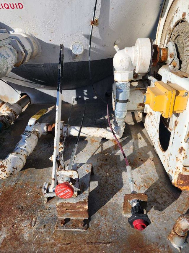 Trucking equipment setup on a metal deck showing hydraulic lines, valve, and control handles near a white tank and rusted components.