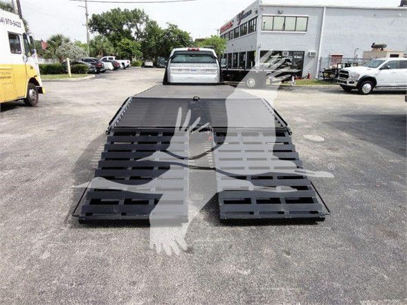 Flatbed tow truck with black modular deck ramps extending rearward, Chevrolet Silverado 6500HD stock truck lot in background.