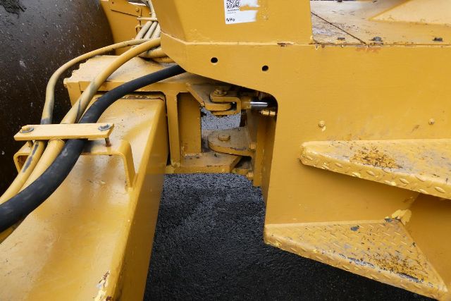 Section of a Bomag BW212D-2 compactor showing yellow metal frame, hydraulic hoses, and a hinge/connection between chassis components.