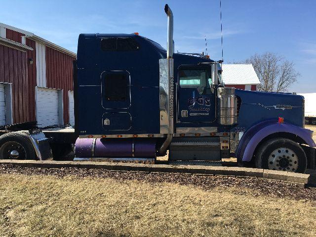 Kenworth W900L truck side view showing blue sleeper cab, chrome exhaust stacks, fuel tanks, and purple accents.
