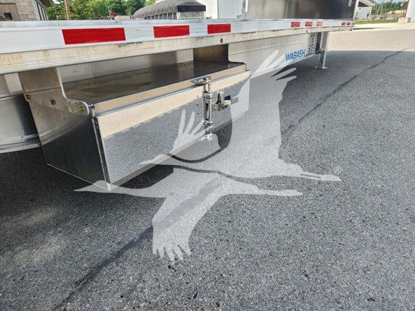 Wabash drop deck trailer undercarriage showing storage toolbox and stabilizer near rear, chrome finish with reflective red/white safety rails.
