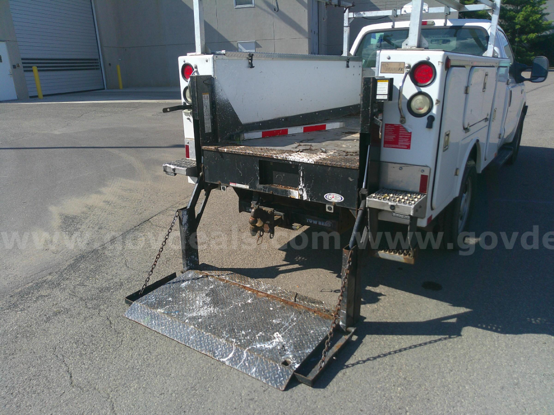 Truck bed with hydraulic or manual lift and folding tailgate on a white Ford utility truck. Diamond plate ramp lowered, chains and steps visible. Rear view shows tail lights and side compartments.