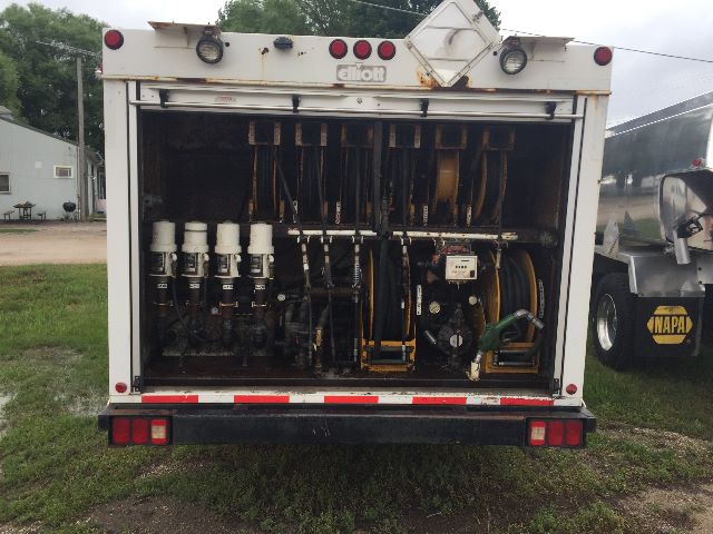 Back of a Freightliner service body showing multiple hydraulic filter housings and hoses, engine components, and pump assembly in the cargo area.