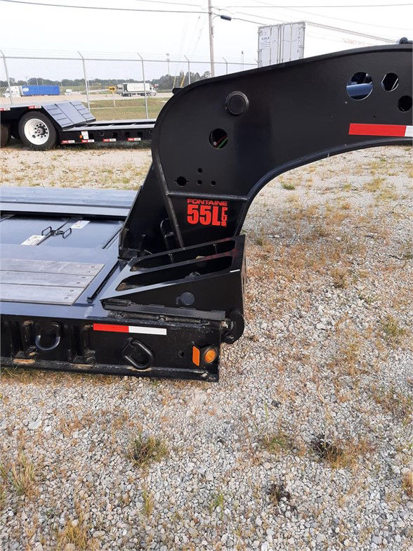 Fontaine 55,000 lb lowboy trailer front corner showing black frame with red ‘55L’ logo and rear deck edge; gravel lot in background.