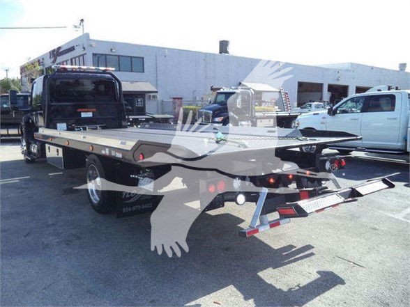 Flatbed tow truck with a black cab and white International MV truck in the lot. Visible text on stickers/branding on the flatbed not clearly legible.