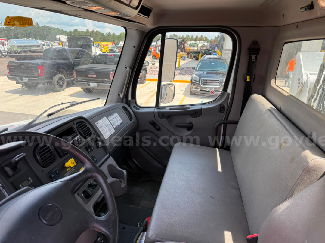 Truck cab interior showing steering wheel, dashboard, gray bench seat, driver-side door panel, and windshield with outdoor lot in background.