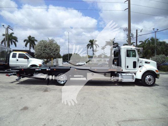 2018 Peterbilt 337 tow truck chassis with a white cab and long flatbed trailer in a lot. Visible text includes Peterbilt badge and standard truck components.