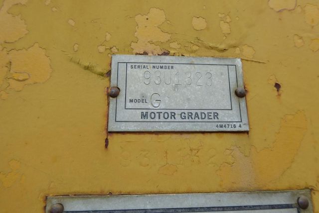 Serial number plate on yellow machinery: 'Serial Number 9301323' and 'Model G' with 'Motor Grader' and '4M04716 4' markings.