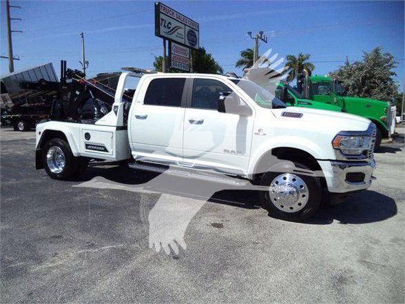 Ram 5500 tow truck side view with white crew cab and long wheelbase, stickered logos, chrome wheels, and equipment bed behind cab.
