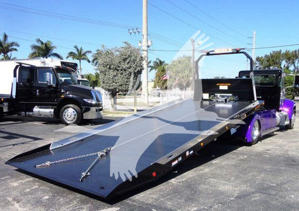 Flatbed tow truck with a purple cab in the background; black equipment on the bed; visible text: 'PETERBILT' on the front of the cab? and general tow truck components.