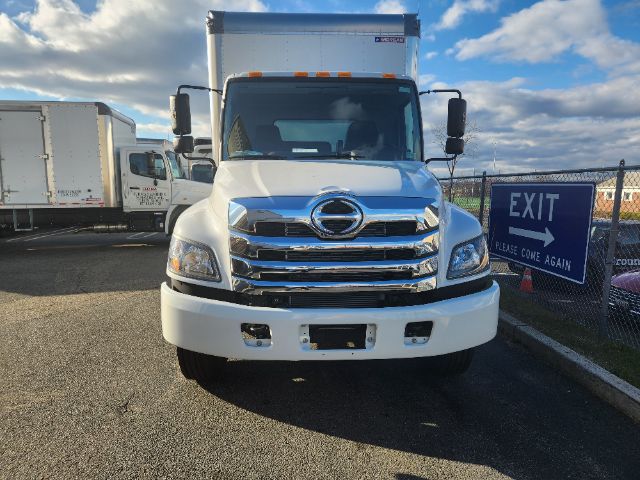 Front view of a white 2025 Hino L6 box truck with chrome grille and bumper, black mirrors, and cargo box against a blue sky.