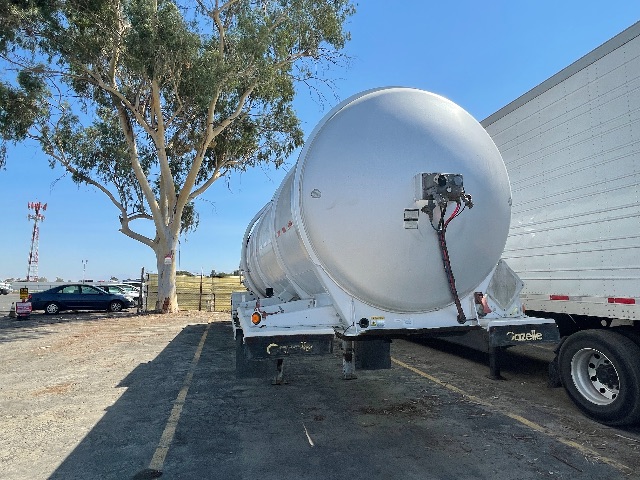 2011 Heil 407 trailer with a cylindrical tank mounted on a dual-axle trailer. The tank has rear entry and hoses, with a white cab and a neighboring white trailer. Clear sky and sparse trees.