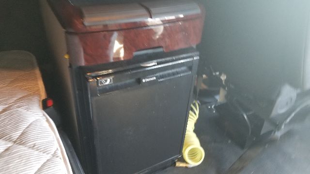 Cab interior of a 2013 Freightliner Coronado 132 showing a small black refrigerator/freezer unit and a yellow hose in a sleeper area.
