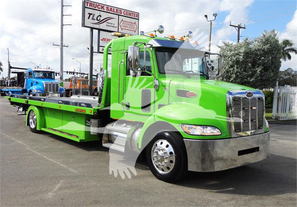 Green Peterbilt tow truck with a long flatbed, chrome bumper, and dual side mirrors. Visible signs include a TLC Truck Sales Center banner in the background.