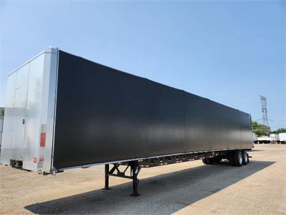Manac COMBO flatbed trailer in-service viewed from side showing long black side panels and silver front corner; trailer on landing gear over gravel.
