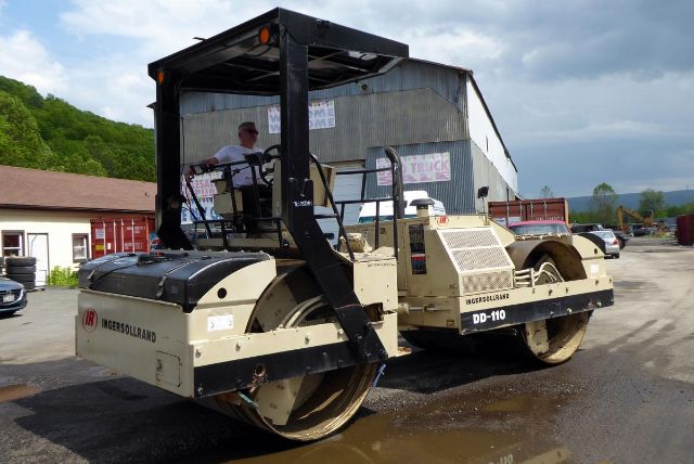 Ingersoll-Rand DD110 pneumatic roller in an outdoor yard, beige with black frame, operator in cabin, large steel drums, Towable rolling compactor.